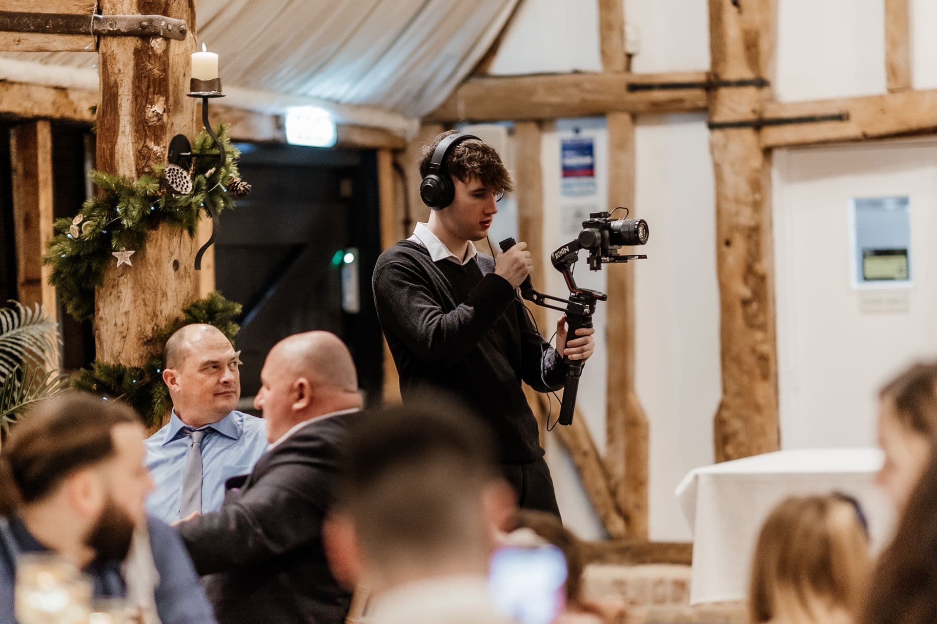 Videographer with headphones films an event using a gimbal camera in a rustic barn.