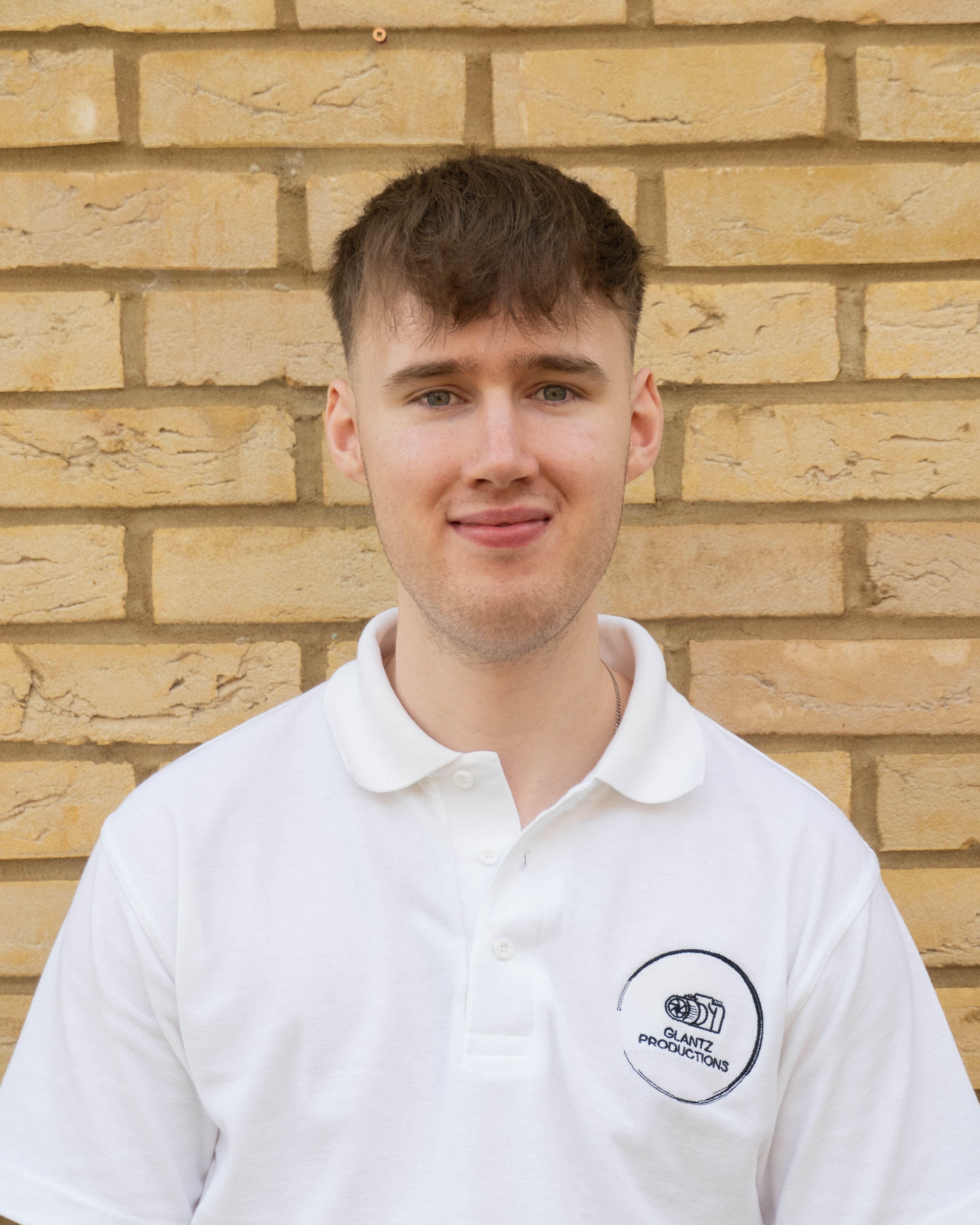 Smiling young man in a white Glantz Productions polo shirt before a brick wall.