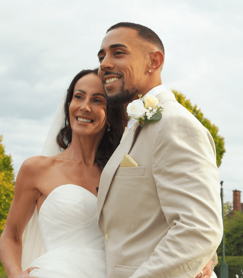 Smiling bride in white strapless gown and groom in beige suit posing outdoors together.