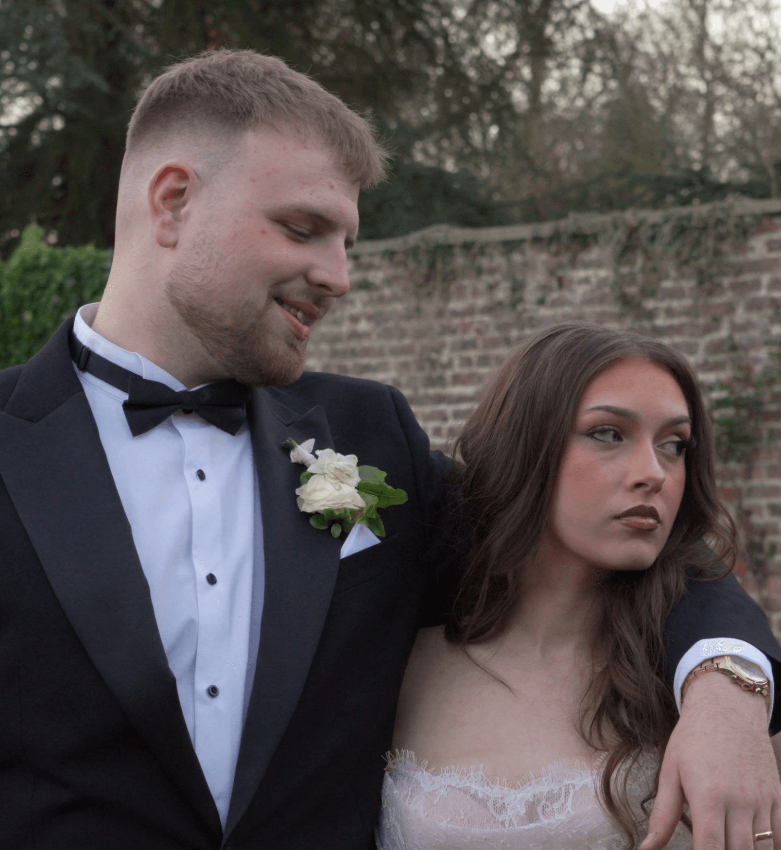 Smiling man in a tuxedo looks at a serious woman in a white lace dress.