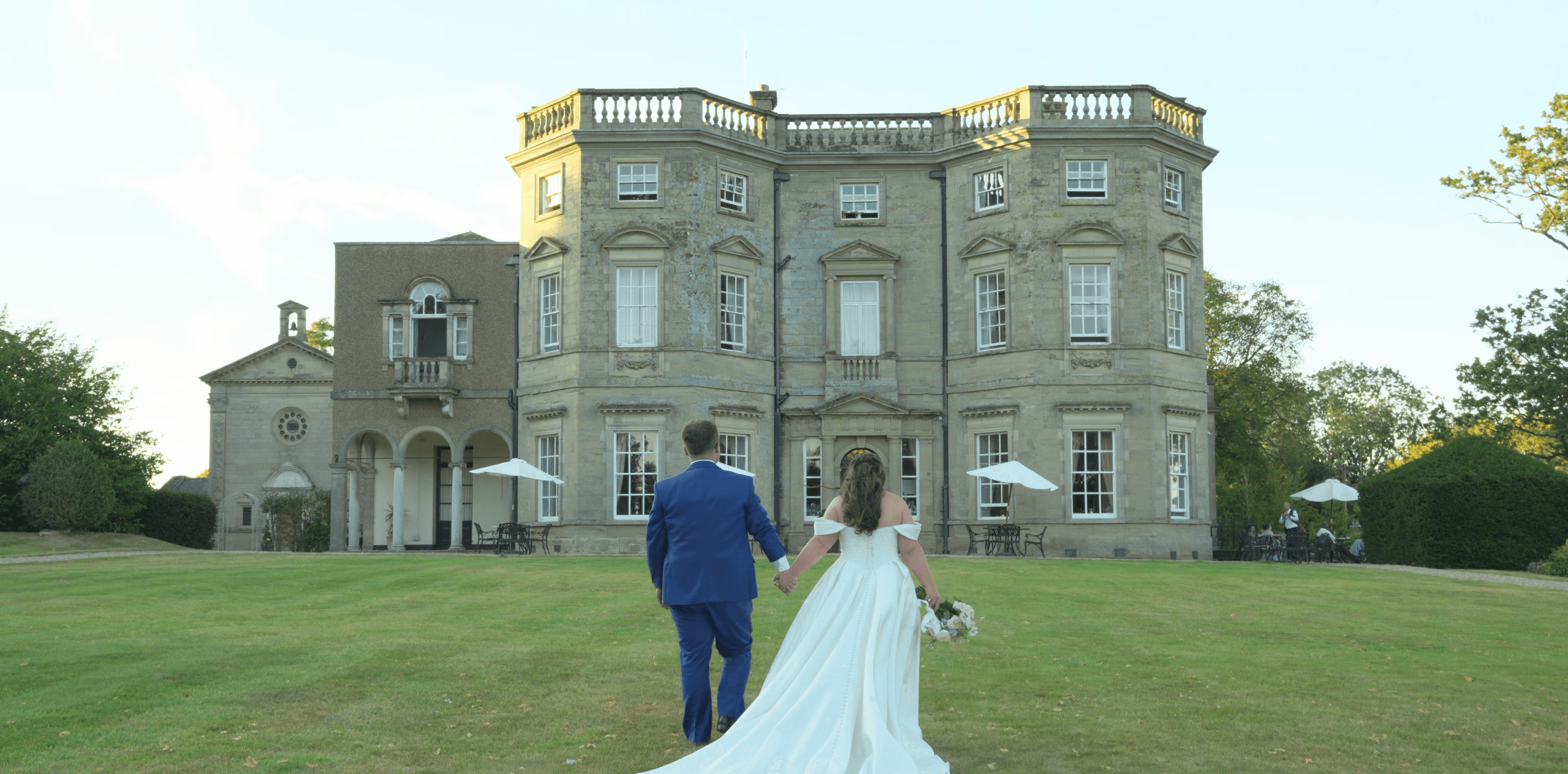 A bride and groom walk hand-in-hand across a green lawn toward a grand stone manor.