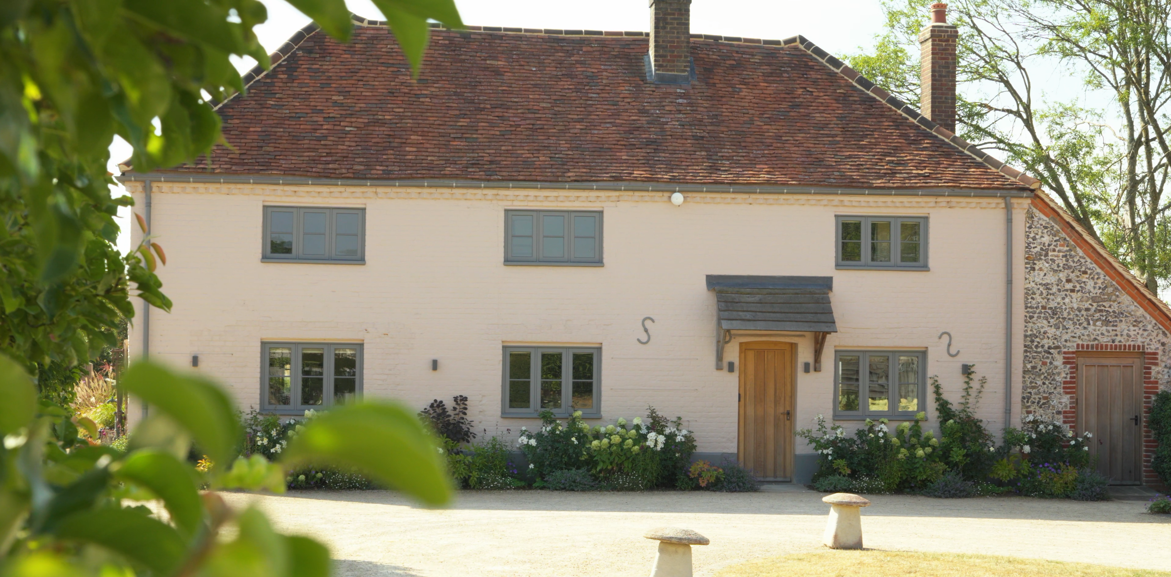 Traditional pink brick house with grey windows, red tiled roof, and white flowers in front.