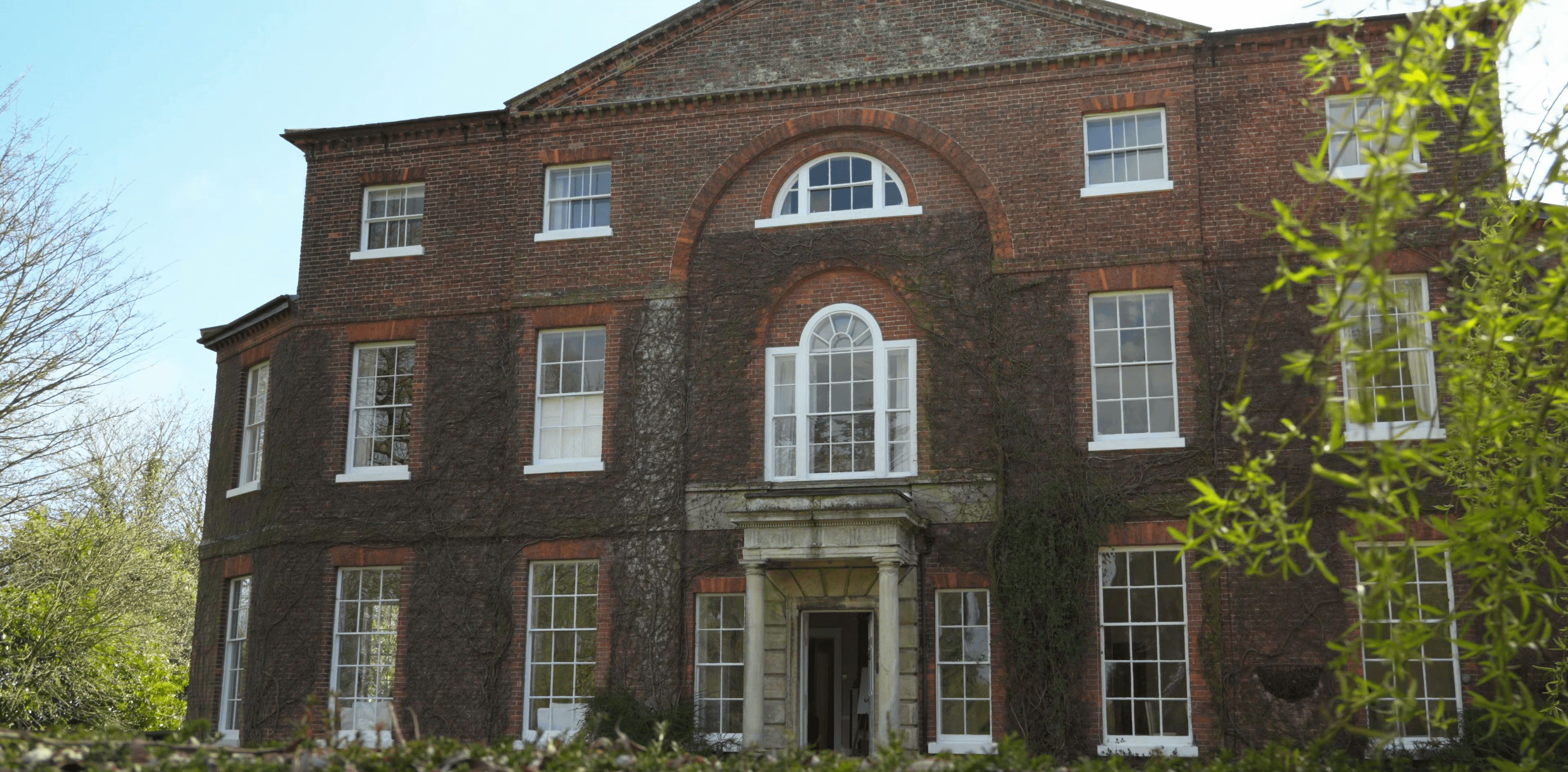 Red brick manor house with white sash windows, climbing ivy, and a stone portico entrance.