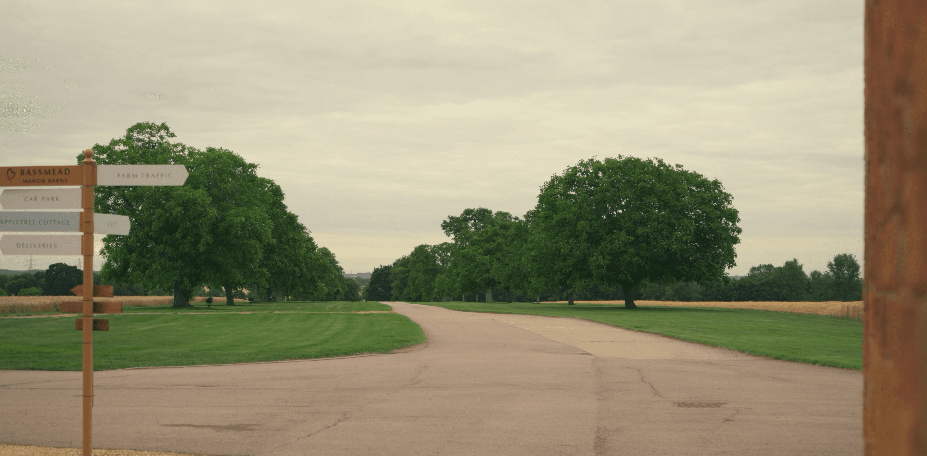 A paved road winds through green lawns and leafy trees past a wooden directional signpost.