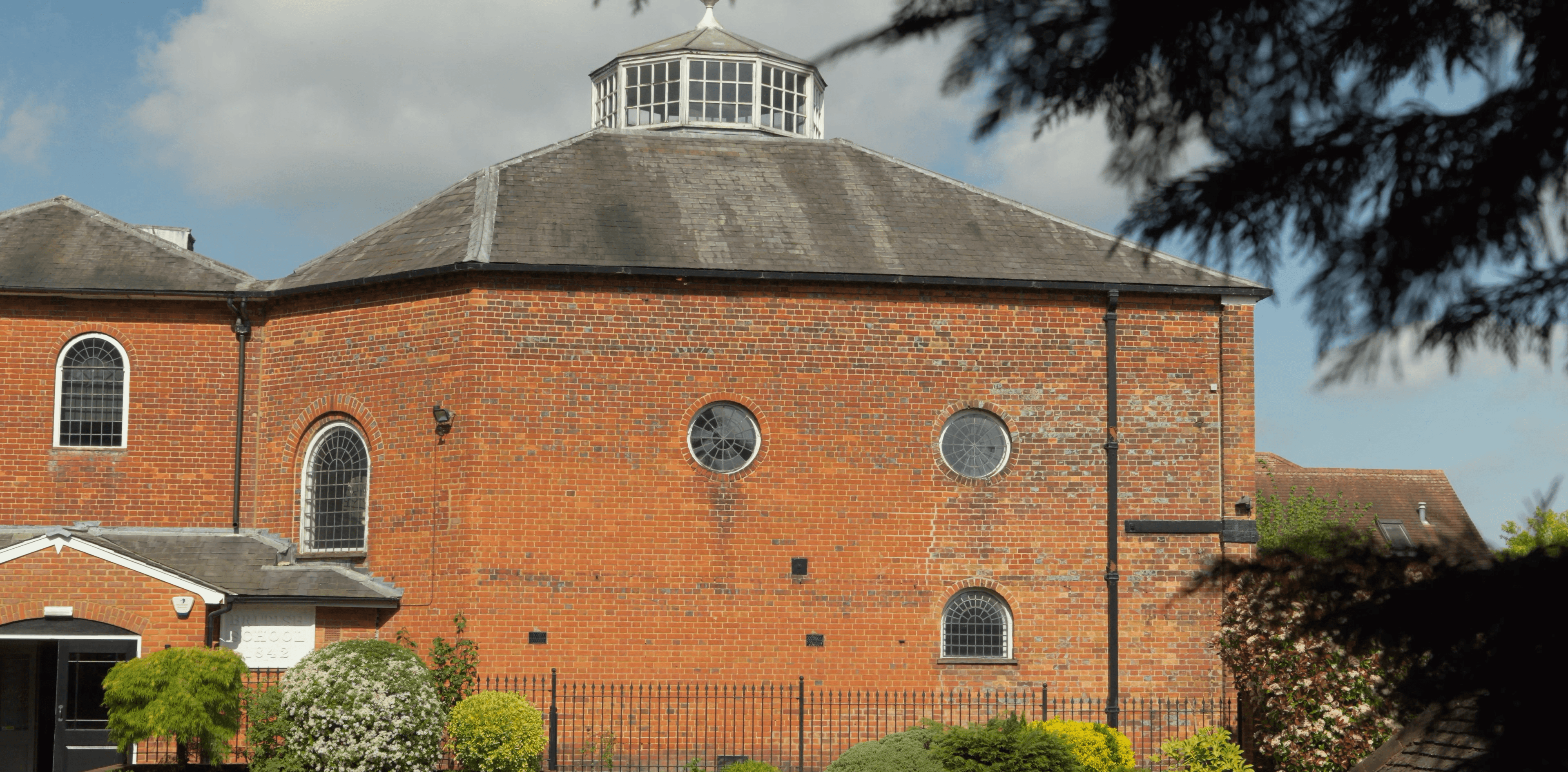 Red brick octagonal building with a white cupola, grey roof, and arched windows.