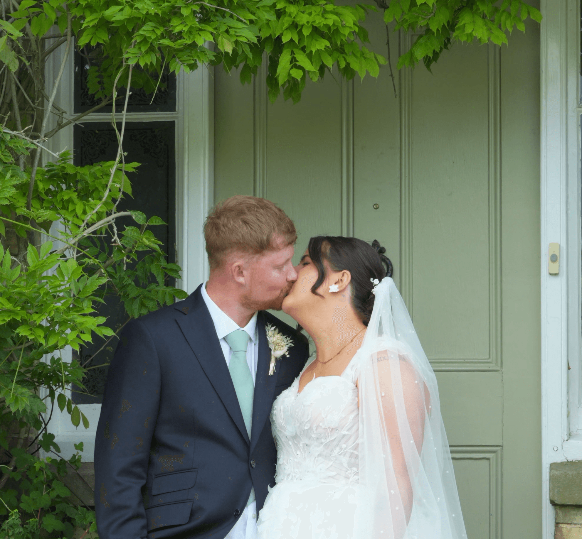 Newlyweds kissing in front of a sage green door, framed by lush green leaves.