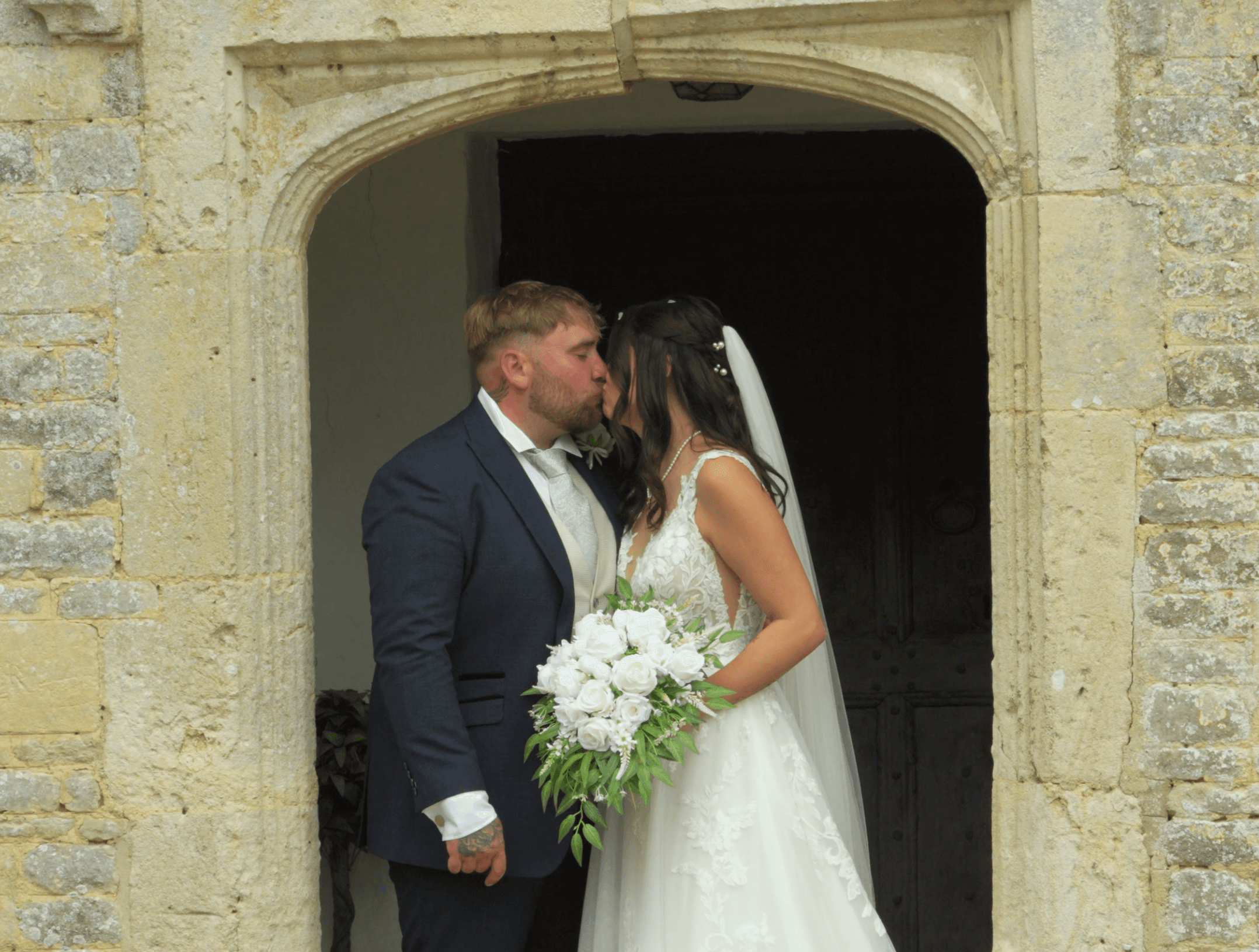 Bride and groom kissing in a stone archway, bride holding a white rose bouquet.