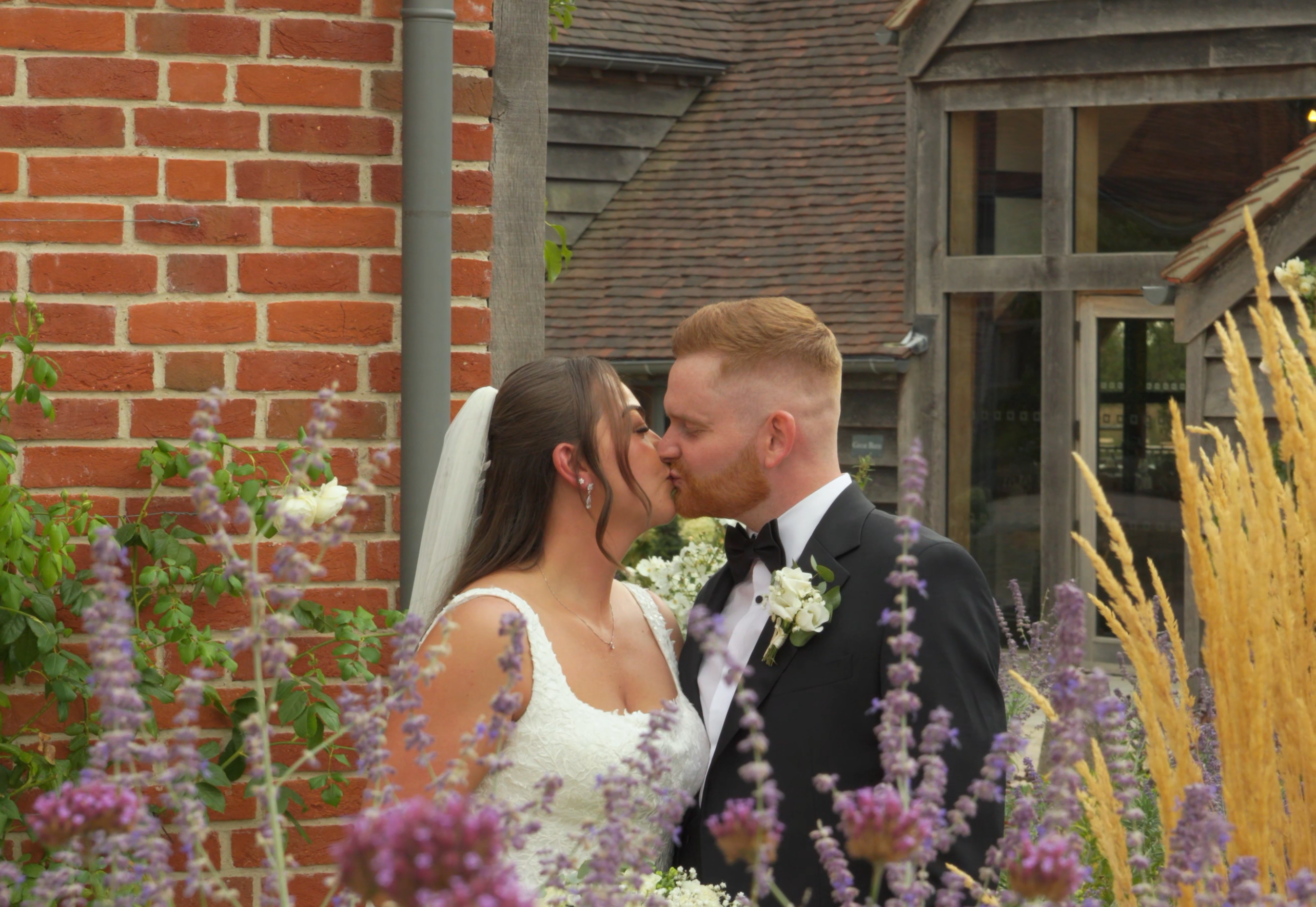 A bride and groom share a romantic kiss behind purple flowers in a garden setting.