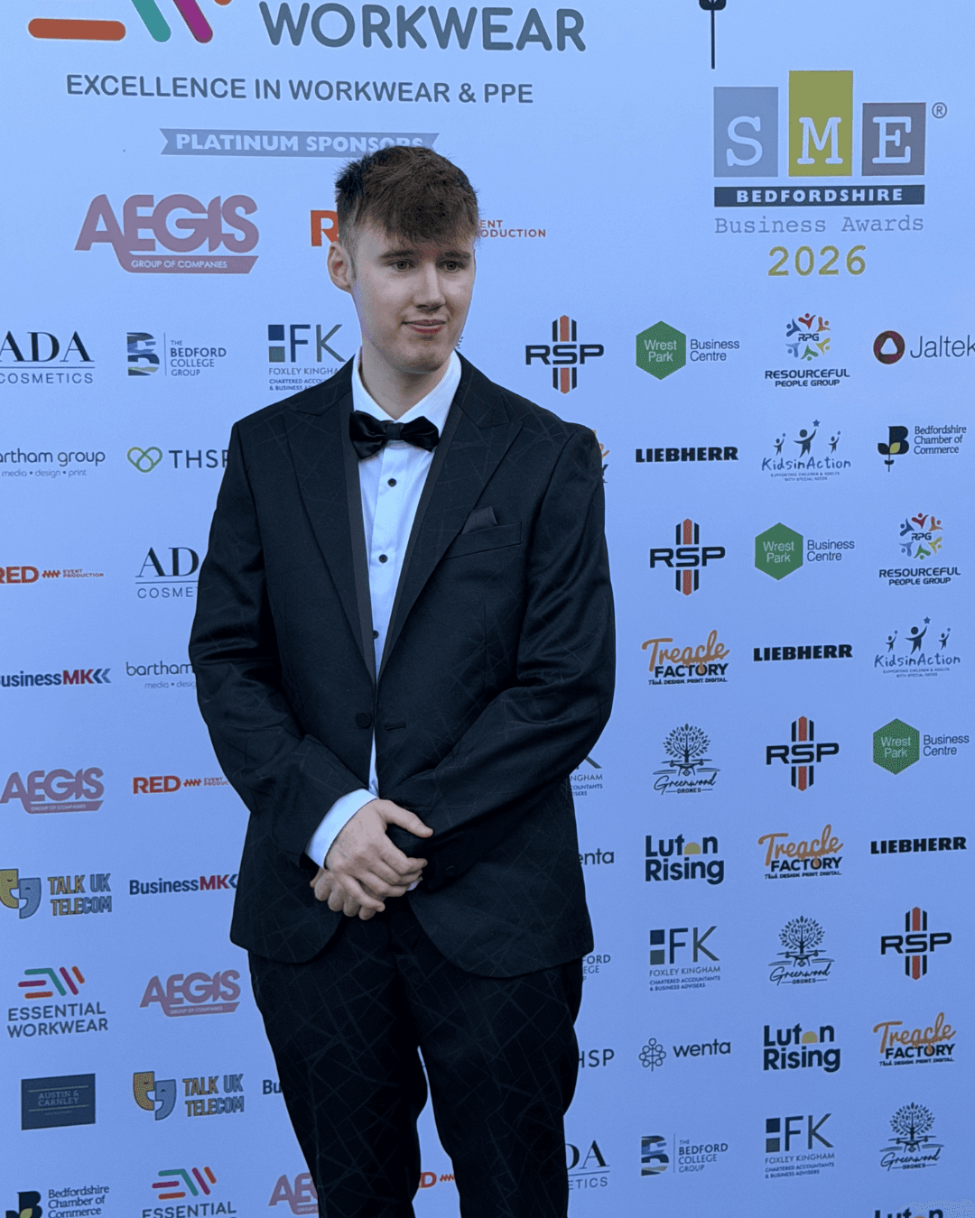 Young man in a black tuxedo and bowtie posing against a branded business awards backdrop.