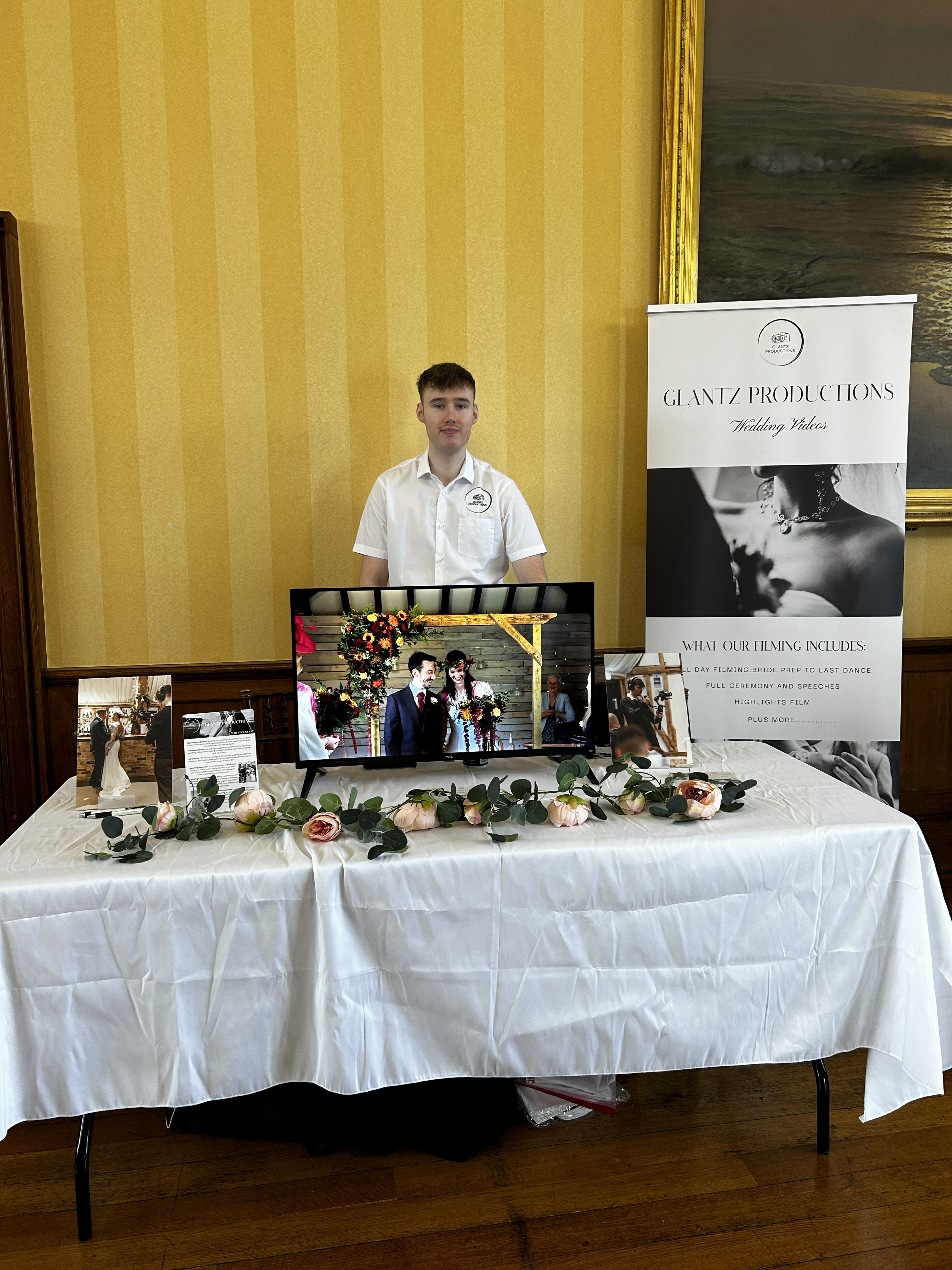 Man standing behind a wedding videography display table with a TV and promotional banner.